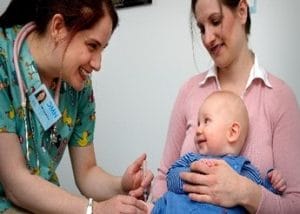 MRT (7by5) A smiling nurse prepares to give a vaccination to a baby sitting on their mother’s lap. The baby looks up at the mother, who is smiling reassuringly.