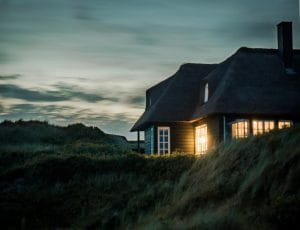 A cozy house with a thatched roof glows warmly at dusk, nestled among tall grass and rolling hills under a cloudy evening sky.