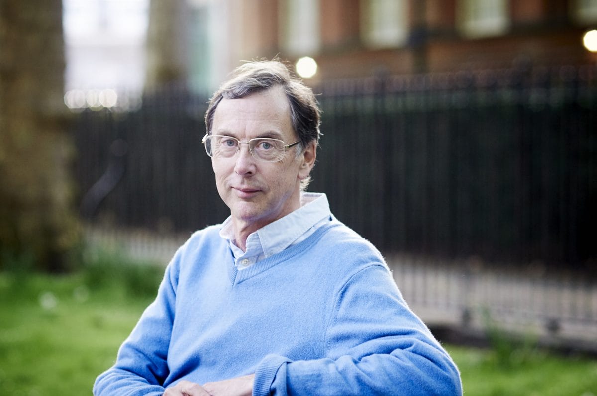 A middle-aged man wearing glasses and a light blue sweater sits outdoors on grass, with a black iron fence and brick building blurred in the background.