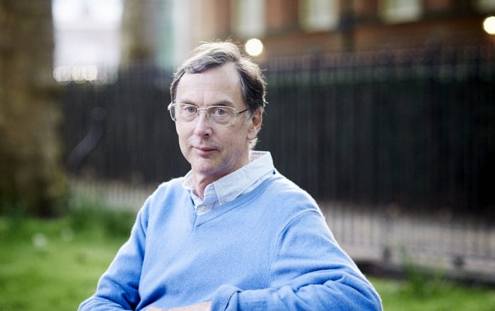 A middle-aged man wearing glasses and a light blue sweater sits outdoors on grass, with a black iron fence and brick building blurred in the background.