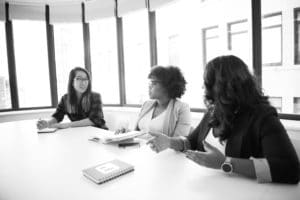 Black and White. 3 Women. Photo by Christina Morillo from Pexels Three women sit at a conference table in a bright office, engaged in conversation. One woman has a notebook open, while the others gesture as they speak, suggesting a collaborative meeting or discussion.