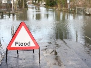 A triangular warning sign reading Flood stands on a road that is heavily covered in water, indicating flooding. Trees and a fence line the sides of the submerged road.