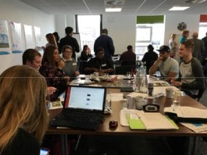 A group of people sit around tables in a bright meeting room, talking and working with laptops, papers, and drinks. Some are engaged in conversation, while others focus on their devices or notes.