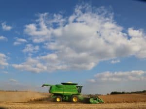 GLOBAL~2 A green combine harvester moves through a golden wheat field under a blue sky with scattered white clouds, kicking up dust as it harvests crops.