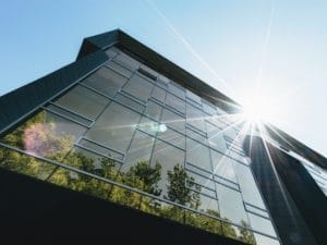 LCCC 4×3 Low-angle view of a modern glass building reflecting green trees, with sunlight shining brightly in the clear blue sky, creating a starburst effect near the roof.