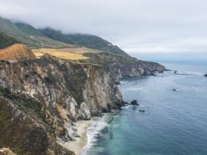 LWEC 4×3 Rocky coastal cliffs border the ocean with waves crashing below, while green hills rise up in the background under a cloudy, misty sky.