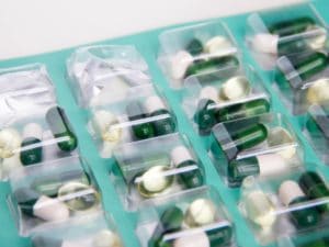 Close-up of a blister pack containing various capsules and softgel pills, including green, white, and transparent varieties, arranged in individual compartments against a teal background.