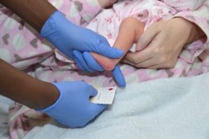 A healthcare worker in blue gloves collects a blood sample from a baby’s heel onto a card, while an adult gently holds the baby, who is dressed in pink pajamas with heart patterns.
