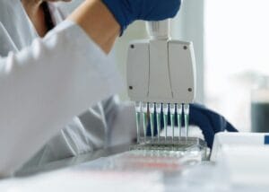A person wearing blue gloves uses a multi-channel pipette to dispense liquid into a microplate in a laboratory setting.