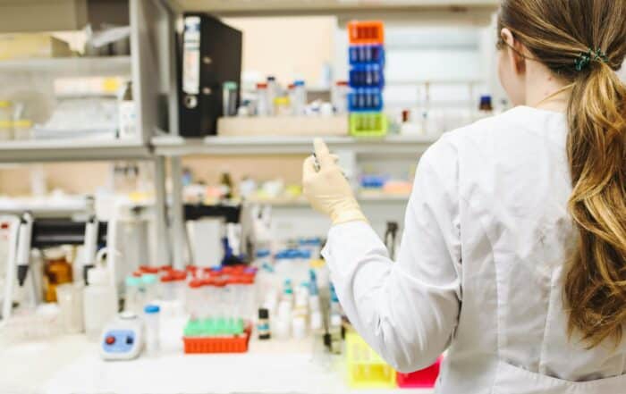 A person in a white lab coat and gloves stands at a laboratory bench, holding a pipette, surrounded by test tubes, racks, and various lab equipment. Shelves with bottles and supplies are visible in the background.