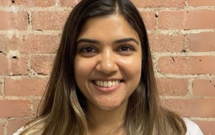 A woman with long, straight brown hair and a white shirt smiles in front of a light brown brick wall.