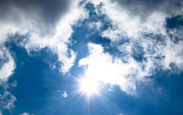Bright sun shining in a blue sky, partially obscured by fluffy white clouds, with sun rays radiating outward and patches of blue visible between the clouds.