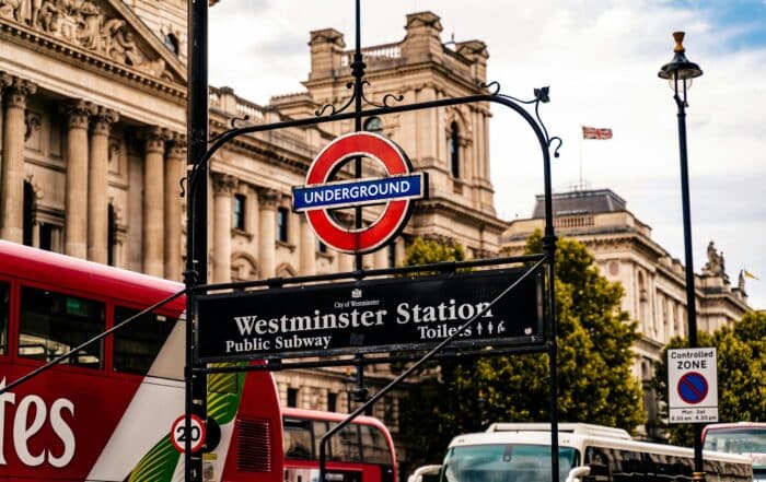 Sign for Westminster Station on the London Underground, with classic red and blue roundel, in front of historic buildings and a red double-decker bus; trees and a cloudy sky in the background.