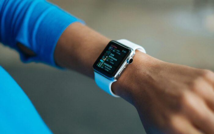 A person wearing a blue long-sleeve shirt checks a silver smartwatch with a white band on their wrist. The smartwatch screen displays various colorful app icons and information.