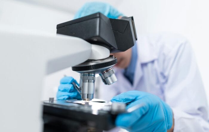 A scientist wearing blue gloves and a lab coat examines samples under a microscope in a laboratory setting.