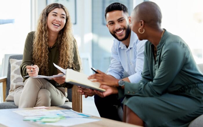 Three people sit together smiling and talking, holding notebooks and papers, suggesting a friendly, collaborative meeting or discussion in a bright, modern office setting.