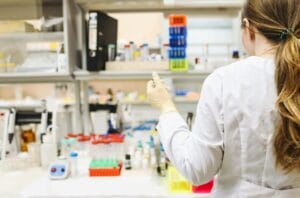 Scientist in a lab A female scientist in a white lab coat and gloves holds a test tube while working in a laboratory filled with lab equipment, test tubes, and bottles on the counter.