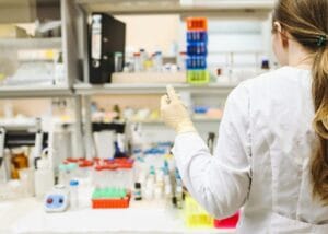 A female scientist in a white lab coat and gloves holds a test tube while working in a laboratory filled with lab equipment, test tubes, and bottles on the counter.