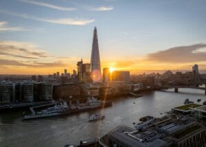 A cityscape of London at sunset, featuring the Shard skyscraper, the River Thames, and boats on the water, with sunlight reflecting off the buildings and river.