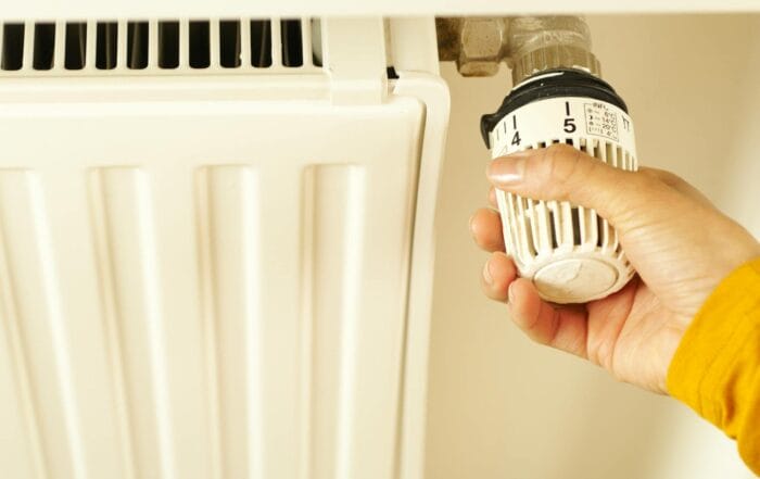 A person’s hand adjusts the temperature dial on a white radiator, turning the knob to regulate heating.