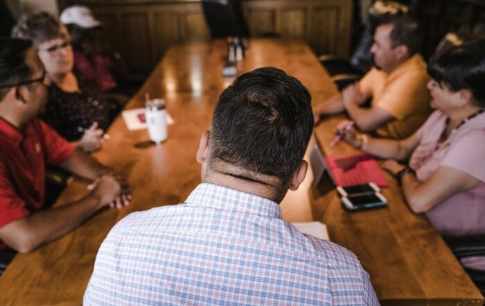 A group of six people sit around a large wooden conference table, engaged in discussion. The photo is taken from behind one man in a checkered shirt, facing the others. Notebooks and drinks are on the table.