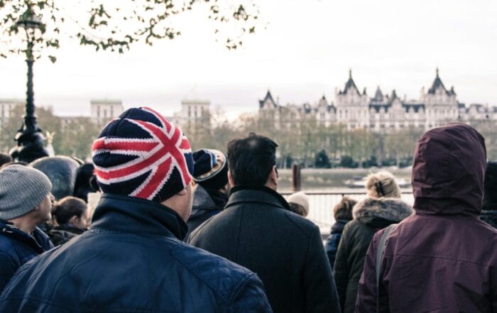 A crowd stands outdoors facing a historic building across a river. One person in the foreground wears a winter hat with the Union Jack design. The weather appears cold and overcast.