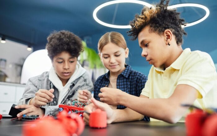 Three kids at school sit at a table working together on an electronics project, handling wires and components, with a circular light fixture above them.