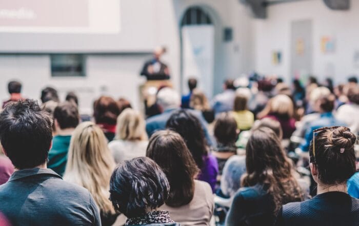 A group of people sit in rows facing a speaker at the front of a bright, modern room, listening to a presentation. The speaker and screen are out of focus, emphasizing the audience.