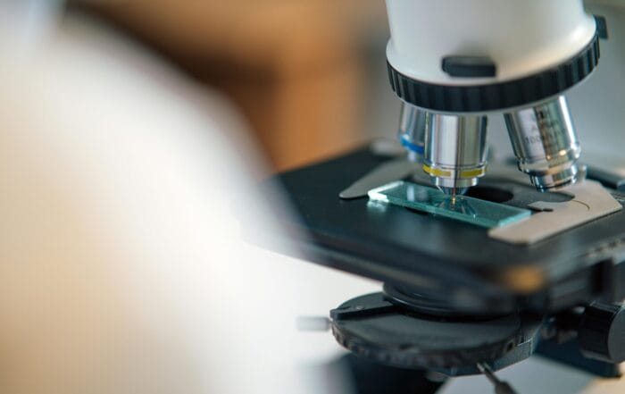 Close-up of a microscope with a glass slide placed on its stage, showing the objective lenses and part of the focusing mechanism in a laboratory setting.