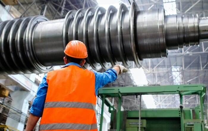 A worker in an orange safety vest and helmet points at large industrial machinery inside a spacious factory with a high, metal-framed ceiling.