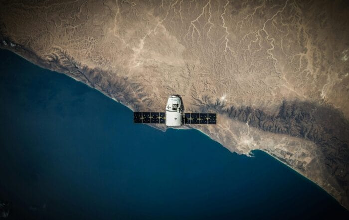 A satellite with solar panels orbits above a coastal landscape, showing a sharp contrast between the deep blue sea and the brown, arid land with visible mountain ridges and winding patterns.
