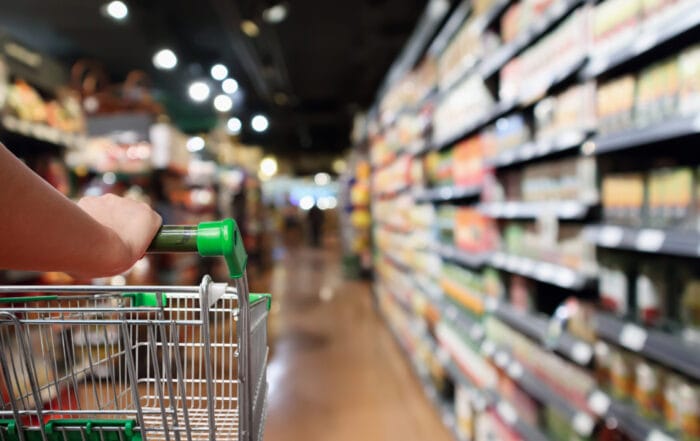 A person pushes a shopping cart down an aisle in a brightly lit supermarket, with shelves stocked with various products visible on the right side of the image.