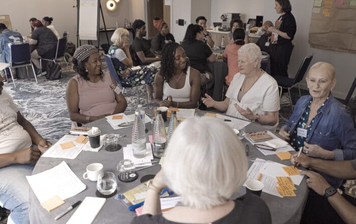 A diverse group of people sit around a round table, engaged in lively discussion, with papers, pens, and drinks in front of them. Other groups are visible in the background, working and talking in a bright, modern room.