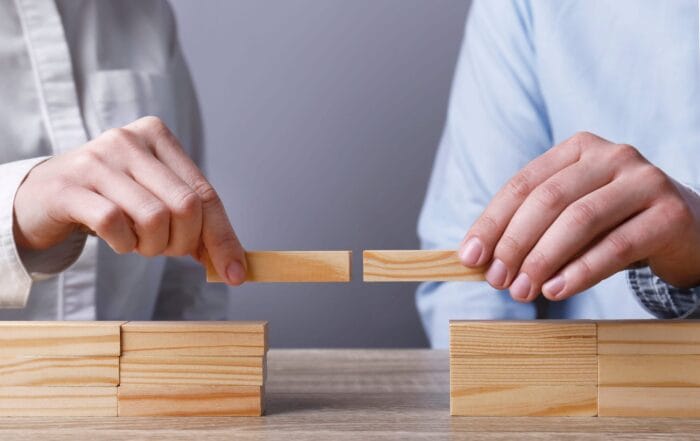 Two people each hold a wooden block, joining them together to bridge a gap between two stacks of wooden blocks on a table, symbolizing connection or collaboration.