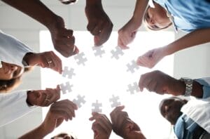 Untitled design (30) A group of people standing in a circle, each holding a white puzzle piece up toward the center, symbolizing teamwork and collaboration. The image is taken from below with bright light in the background.
