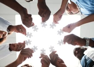 A group of people standing in a circle, each holding a white puzzle piece up toward the center, symbolizing teamwork and collaboration. The image is taken from below with bright light in the background.