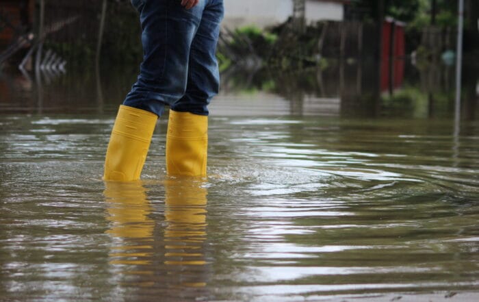 A person wearing yellow rubber boots and jeans stands in deep floodwater outdoors, with water reflecting the surroundings and some buildings and a fence visible in the background.