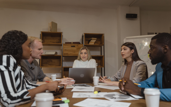 Five people sit around a table in an office, having a discussion with documents, pens, and coffee cups in front of them. One person has a laptop open, and there are shelves and a whiteboard in the background.