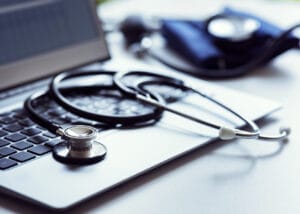 A stethoscope rests on a laptop keyboard, with a blood pressure cuff in the background, symbolizing digital healthcare or telemedicine.