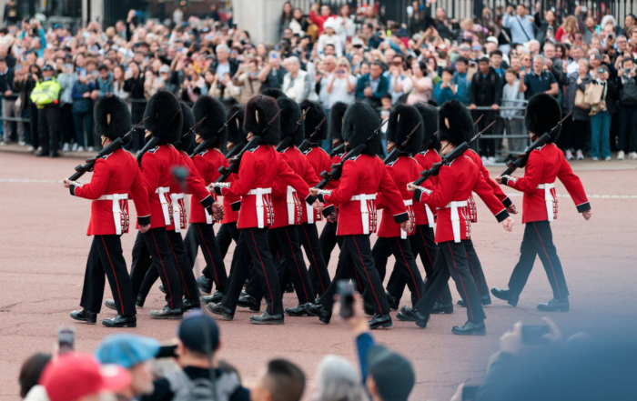 A group of guards in red uniforms and tall black hats march in formation, watched by a large crowd of spectators behind barriers. Some people in the crowd are taking photos.