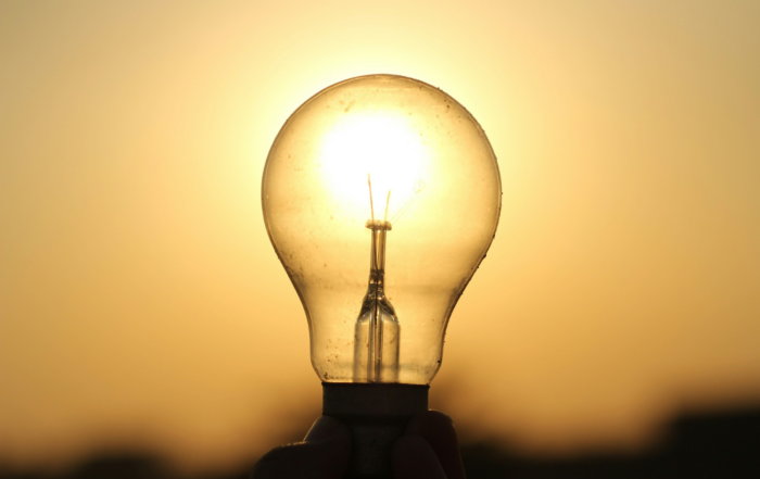 A hand holds a clear light bulb against a bright sunset.